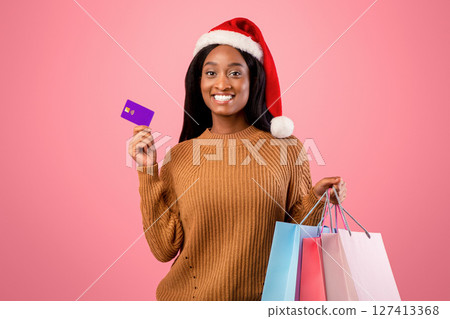 Online Christmas sale concept. Happy black woman in Santa hat holding shopping bags and credit card on pink studio background. Positive African American lady with packed Xmas purchases and bank card 127413368