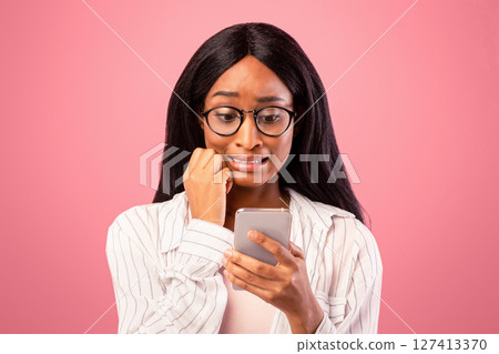 Oops. Embarassed black woman looking at mobile phone screen in confusion on pink studio background. Emotional African American lady making mistake while working in web 127413370