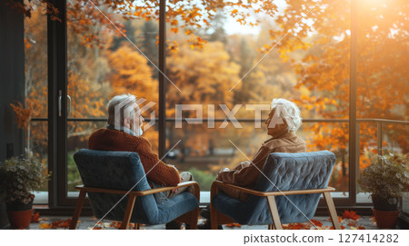 An elderly couple sits by a window, looking out at the fall foliage. They are enjoying a peaceful moment together, surrounded by the beauty of the season. 127414282