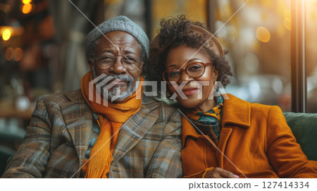 An older couple smiles for a photo at a cafe, the man wearing a gray hat and orange scarf and the woman in a bright orange coat. 127414334
