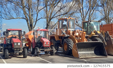 Multiple construction and agriculture vehicles, including tractors and loaders, are parked in an open lot. The scene highlights machinery ready for industrial and farming use Multiple construction and agriculture vehicles, including tractors and loaders, are parked in an open lot. The scene highlights machinery ready for industrial and farming use 127415575
