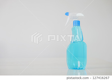 Blue glass spray bottle with white trigger in the kitchen table on a white wall background. The bottle is filled with clear blue liquid, Cleaning concept 127416267