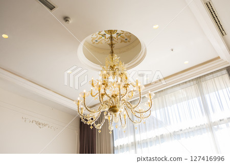 Elegant chandelier hanging from decorated ceiling in luxury hotel interior. Classic design, ornate lighting, and refined architectural style concept. 127416996