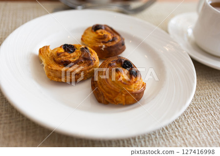 Three raisin pastries served on white plate during hotel breakfast. Morning dessert, sweet bakery snack, and elegant table setting concept. Three raisin pastries served on white plate during hotel breakfast. Morning dessert, sweet bakery snack, and elegant table setting concept. 127416998