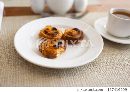 Three raisin pastries served on white plate during hotel breakfast. Morning dessert, sweet bakery snack, and elegant table setting concept. Three raisin pastries served on white plate during hotel breakfast. Morning dessert, sweet bakery snack, and elegant table setting concept. 127416999
