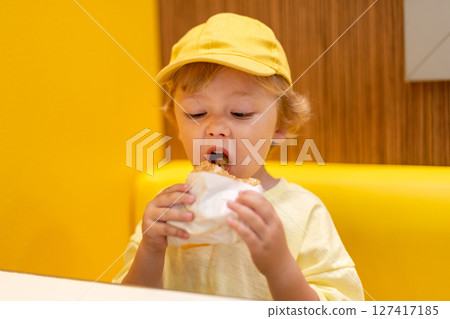 Young child wearing a yellow hat and shirt eats a cheeseburger in a vibrant restaurant, enjoying fast food meal in a casual dining environment. Kids diet, bad food choices, early junk food addiction 127417185