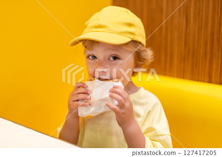 Young child wearing a yellow hat and shirt eats a cheeseburger in a vibrant restaurant, enjoying fast food meal in a casual dining environment. Kids diet, bad food choices, early junk food addiction Young child wearing a yellow hat and shirt eats a cheeseburger in a vibrant restaurant, enjoying fast food meal in a casual dining environment. Kids diet, bad food choices, early junk food addiction 127417205