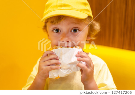 Young child wearing a yellow hat and shirt eats a cheeseburger in a vibrant restaurant, enjoying fast food meal in a casual dining environment. Kids diet, bad food choices, early junk food addiction Young child wearing a yellow hat and shirt eats a cheeseburger in a vibrant restaurant, enjoying fast food meal in a casual dining environment. Kids diet, bad food choices, early junk food addiction 127417206