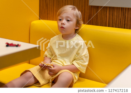 Young child wearing a yellow clothes eats a cheeseburger in a vibrant restaurant, enjoying fast food meal in a casual dining environment. Kids diet, bad food choices, early junk food addiction 127417218
