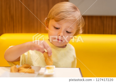A small child wearing a yellow shirt is eating chicken nuggets in a vibrant environment. Four year old boy enjoying fast food meal. Kids diet, bad food choices, early junk food addiction A small child wearing a yellow shirt is eating chicken nuggets in a vibrant environment. Four year old boy enjoying fast food meal. Kids diet, bad food choices, early junk food addiction 127417220