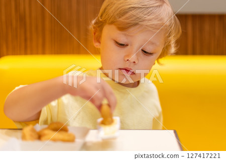 A small child wearing a yellow shirt is eating chicken nuggets in a vibrant environment. Four year old boy enjoying fast food meal. Kids diet, bad food choices, early junk food addiction 127417221