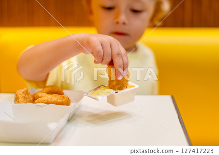 A small child wearing a yellow shirt is eating chicken nuggets in a vibrant environment. Four year old boy enjoying fast food meal. Kids diet, bad food choices, early junk food addiction 127417223