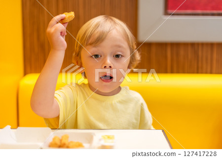 A small child wearing a yellow shirt is eating chicken nuggets in a vibrant environment. Four year old boy enjoying fast food meal. Kids diet, bad food choices, early junk food addiction 127417228