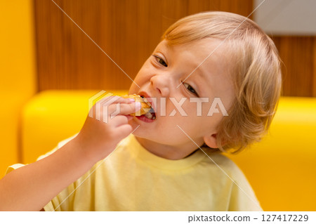 A small child wearing a yellow shirt is eating chicken nuggets in a vibrant environment. Four year old boy enjoying fast food meal. Kids diet, bad food choices, early junk food addiction A small child wearing a yellow shirt is eating chicken nuggets in a vibrant environment. Four year old boy enjoying fast food meal. Kids diet, bad food choices, early junk food addiction 127417229