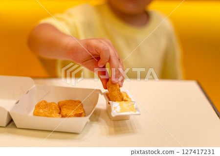 A small child wearing a yellow shirt is eating chicken nuggets in a vibrant environment. Four year old boy enjoying fast food meal. Kids diet, bad food choices, early junk food addiction 127417231