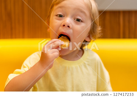 A small child wearing a yellow shirt is eating chicken nuggets in a vibrant environment. Four year old boy enjoying fast food meal. Kids diet, bad food choices, early junk food addiction 127417233
