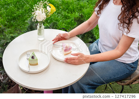 A woman sitting outdoors enjoys vibrant desserts on a white table with a flower vase, surrounded by greenery. The image exudes a serene, relaxing, and indulgent atmosphere. 127417296