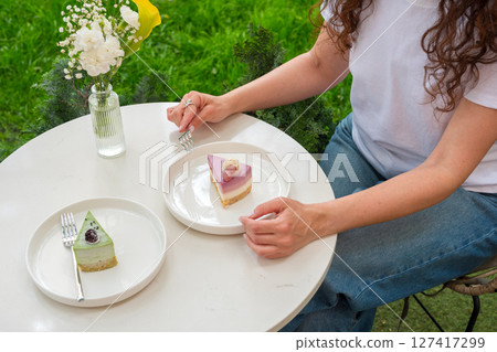 A woman sitting outdoors enjoys vibrant desserts on a white table with a flower vase, surrounded by greenery. The image exudes a serene, relaxing, and indulgent atmosphere. A woman sitting outdoors enjoys vibrant desserts on a white table with a flower vase, surrounded by greenery. The image exudes a serene, relaxing, and indulgent atmosphere. 127417299