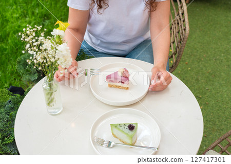 A woman sitting outdoors enjoys vibrant desserts on a white table with a flower vase, surrounded by greenery. The image exudes a serene, relaxing, and indulgent atmosphere. 127417301