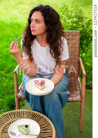 A woman seated at an outdoor table with two plates of desserts, enjoying the scenery and calm surroundings in a peaceful green garden. diet concept, fasting, dieting, losing weight, woman on a diet. A woman seated at an outdoor table with two plates of desserts, enjoying the scenery and calm surroundings in a peaceful green garden. diet concept, fasting, dieting, losing weight, woman on a diet. 127417314