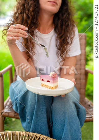 A woman seated at an outdoor table with two plates of desserts, enjoying the scenery and calm surroundings in a peaceful green garden. She smiles thoughtfully, epitomizing comfort and joy in nature. 127417321