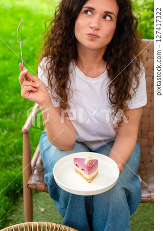 A woman seated at an outdoor table with two plates of desserts, enjoying the scenery and calm surroundings in a peaceful green garden. She smiles thoughtfully, epitomizing comfort and joy in nature. 127417322