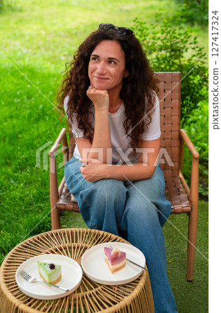 A woman seated at an outdoor table with two plates of desserts, enjoying the scenery and calm surroundings in a peaceful green garden. She smiles thoughtfully, epitomizing comfort and joy in nature. 127417324
