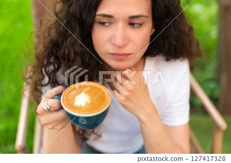 A young woman with curly hair holding a cup of coffee, seated outdoors surrounded by vibrant greenery, radiating a calm and introspective mood in a serene and cozy environment. 127417328