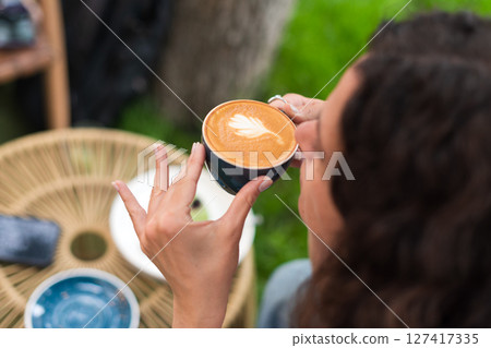 A young woman with curly hair holding a cup of coffee, seated outdoors surrounded by vibrant greenery, radiating a calm and introspective mood in a serene and cozy environment. 127417335