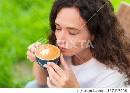 A young woman with curly hair holding a cup of coffee, seated outdoors surrounded by vibrant greenery, radiating a calm and introspective mood in a serene and cozy environment. A young woman with curly hair holding a cup of coffee, seated outdoors surrounded by vibrant greenery, radiating a calm and introspective mood in a serene and cozy environment. 127417338