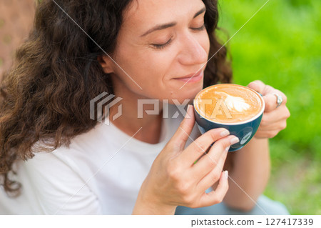 A young woman with curly hair holding a cup of coffee, seated outdoors surrounded by vibrant greenery, radiating a calm and introspective mood in a serene and cozy environment. A young woman with curly hair holding a cup of coffee, seated outdoors surrounded by vibrant greenery, radiating a calm and introspective mood in a serene and cozy environment. 127417339