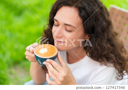 A young woman with curly hair holding a cup of coffee, seated outdoors surrounded by vibrant greenery, radiating a calm and introspective mood in a serene and cozy environment. 127417340