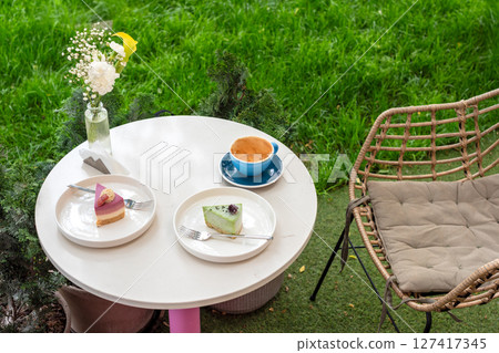 A cozy outdoor caf? table featuring two colorful desserts, a cup of coffee, and a vase of flowers, set against a vibrant garden background, offering a delightful and relaxing atmosphere. A cozy outdoor caf? table featuring two colorful desserts, a cup of coffee, and a vase of flowers, set against a vibrant garden background, offering a delightful and relaxing atmosphere. 127417345