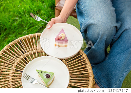 A woman is holding a plate of colorful layered cake while seated in a garden setting. The soft lighting and lush green background enhance the fresh and joyful mood of the scene. 127417347