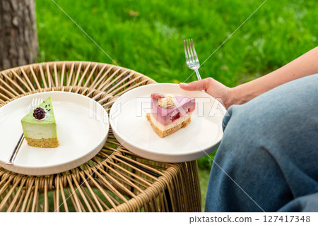 A woman is holding a plate of colorful layered cake while seated in a garden setting. The soft lighting and lush green background enhance the fresh and joyful mood of the scene. 127417348