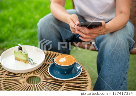 Close-up of a woman holding a cappuccino with latte art and a smartphone during a relaxing outdoors break featuring a plate with a dessert slice on a woven table. Close-up of a woman holding a cappuccino with latte art and a smartphone during a relaxing outdoors break featuring a plate with a dessert slice on a woven table. 127417354