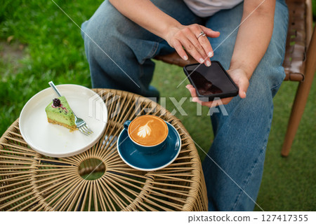 Close-up of a woman holding a cappuccino with latte art and a smartphone during a relaxing outdoors break featuring a plate with a dessert slice on a woven table. Close-up of a woman holding a cappuccino with latte art and a smartphone during a relaxing outdoors break featuring a plate with a dessert slice on a woven table. 127417355