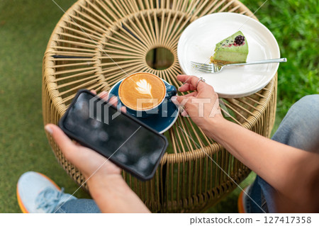Close-up of a woman holding a cappuccino with latte art and a smartphone during a relaxing outdoors break featuring a plate with a dessert slice on a woven table. Close-up of a woman holding a cappuccino with latte art and a smartphone during a relaxing outdoors break featuring a plate with a dessert slice on a woven table. 127417358