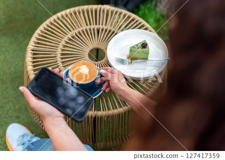 Close-up of a woman holding a cappuccino with latte art and a smartphone during a relaxing outdoors break featuring a plate with a dessert slice on a woven table. Close-up of a woman holding a cappuccino with latte art and a smartphone during a relaxing outdoors break featuring a plate with a dessert slice on a woven table. 127417359