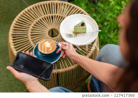 Close-up of a woman holding a cappuccino with latte art and a smartphone during a relaxing outdoors break featuring a plate with a dessert slice on a woven table. Close-up of a woman holding a cappuccino with latte art and a smartphone during a relaxing outdoors break featuring a plate with a dessert slice on a woven table. 127417360