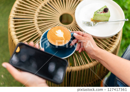 Close-up of a woman holding a cappuccino with latte art and a smartphone during a relaxing outdoors break featuring a plate with a dessert slice on a woven table. 127417361