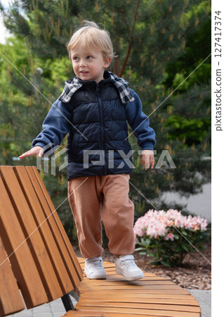 A young child balancing on a curved wooden bench amidst greenery and flowers on a bright day. The scene captures carefree moments in a park. The child is dressed warmly and smiling. 127417374