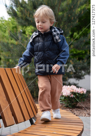 A young child balancing on a curved wooden bench amidst greenery and flowers on a bright day. The scene captures carefree moments in a park. The child is dressed warmly and smiling. 127417375