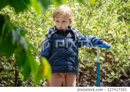 A photo of a young boy dressed for cool weather, holding a scooter, and standing on a pedestrian path surrounded by greenery, capturing a moment of outdoor leisure and exploration. 127417381