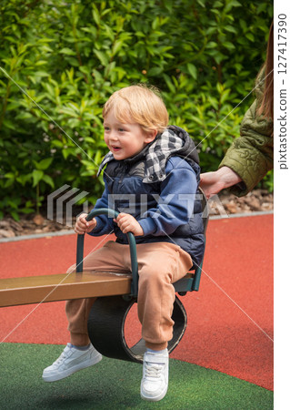Child enjoying a seesaw ride in a sunny outdoor playground area. The setting includes nature surrounding a colorful playground, depicting joy and bonding during leisure time. Child enjoying a seesaw ride in a sunny outdoor playground area. The setting includes nature surrounding a colorful playground, depicting joy and bonding during leisure time. 127417390