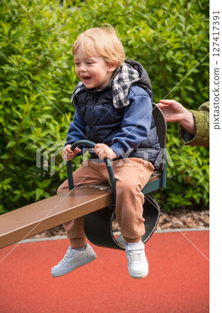 Child enjoying a seesaw ride in a sunny outdoor playground area. The setting includes nature surrounding a colorful playground, depicting joy and bonding during leisure time. 127417391