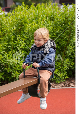 Child enjoying a seesaw ride in a sunny outdoor playground area. The setting includes nature surrounding a colorful playground, depicting joy and bonding during leisure time. Child enjoying a seesaw ride in a sunny outdoor playground area. The setting includes nature surrounding a colorful playground, depicting joy and bonding during leisure time. 127417392