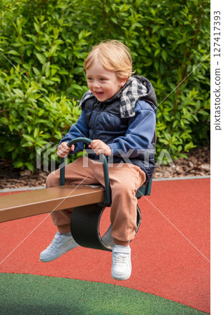 Child enjoying a seesaw ride in a sunny outdoor playground area. The setting includes nature surrounding a colorful playground, depicting joy and bonding during leisure time. Child enjoying a seesaw ride in a sunny outdoor playground area. The setting includes nature surrounding a colorful playground, depicting joy and bonding during leisure time. 127417393