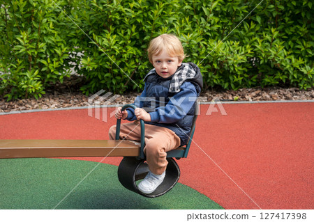 Child enjoying a seesaw ride in a sunny outdoor playground area. The setting includes nature surrounding a colorful playground, depicting joy and bonding during leisure time. Child enjoying a seesaw ride in a sunny outdoor playground area. The setting includes nature surrounding a colorful playground, depicting joy and bonding during leisure time. 127417398