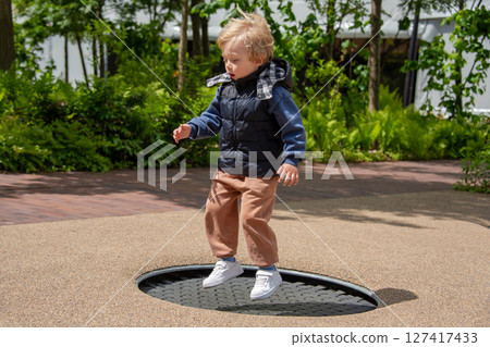 A young child jumping on a trampoline at an outdoor park surrounded by greenery. The child shows excitement and joy while playing, embodying youthful energy and fun in the sunlit environment. 127417433